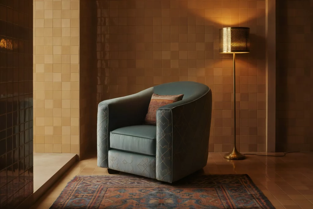 A stylish blue armchair with an intricate pattern, positioned in front of a textured beige tiled wall, accompanied by a vintage lamp and a patterned rug.