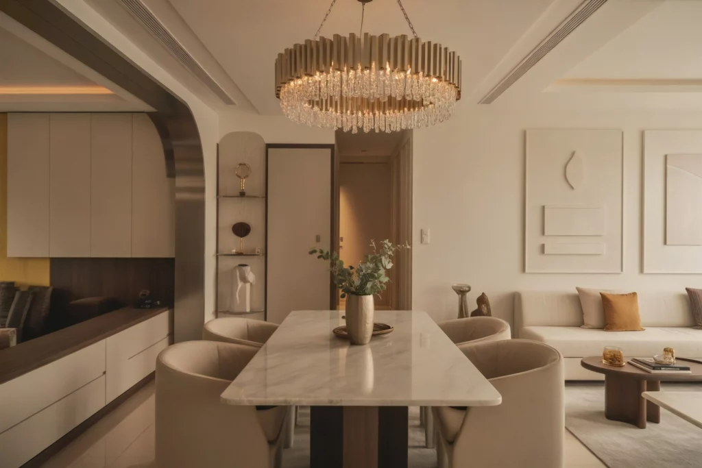 A modern dining area with a sleek marble table, contemporary chairs, and a warm ambiance created by a large chandelier. The space features soft lighting, neutral tones, and minimalist decor, blending elegance with functionality.