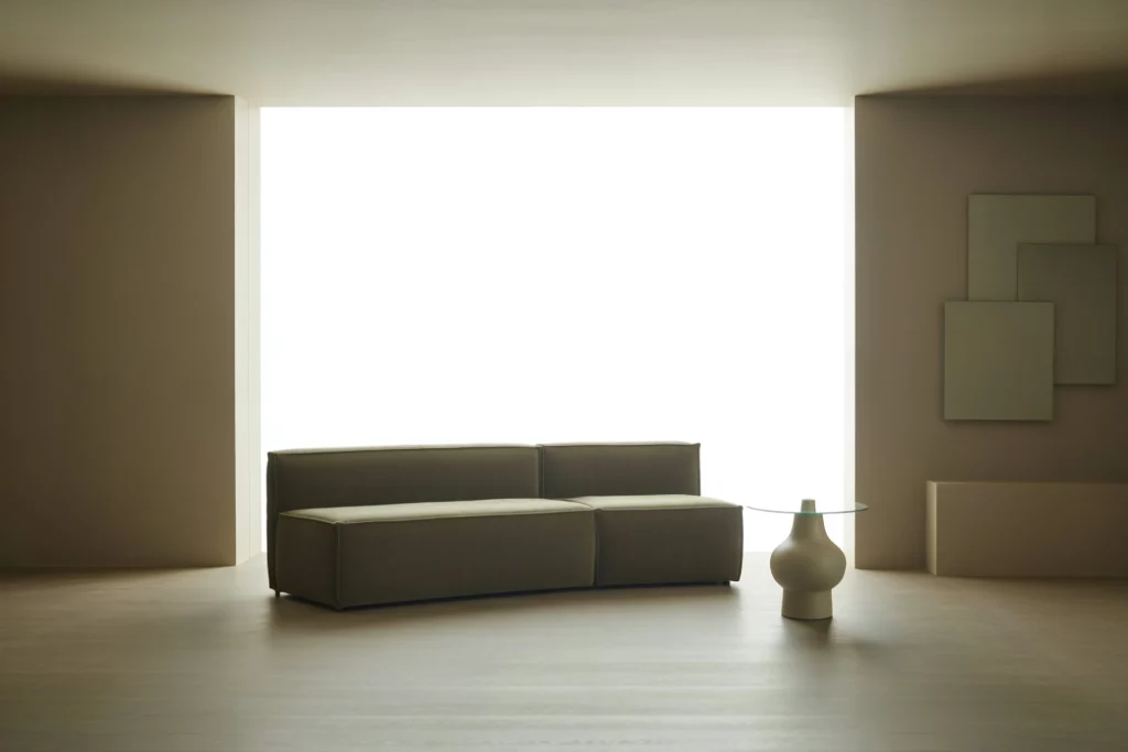 Minimalist living room with neutral tones, featuring a modern brown sofa, glass side table, and abstract wall art.