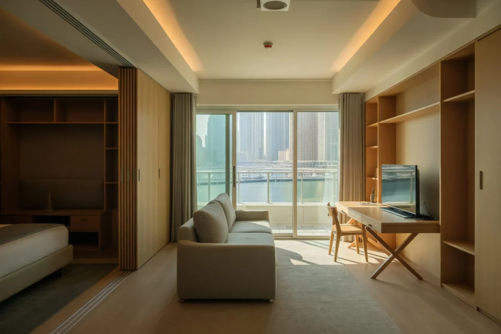 A modern, well-lit apartment with a cozy living area featuring a white sofa, wooden furniture, and built-in storage. Large windows offer a scenic view of the city skyline and waterfront, creating a bright and inviting space.