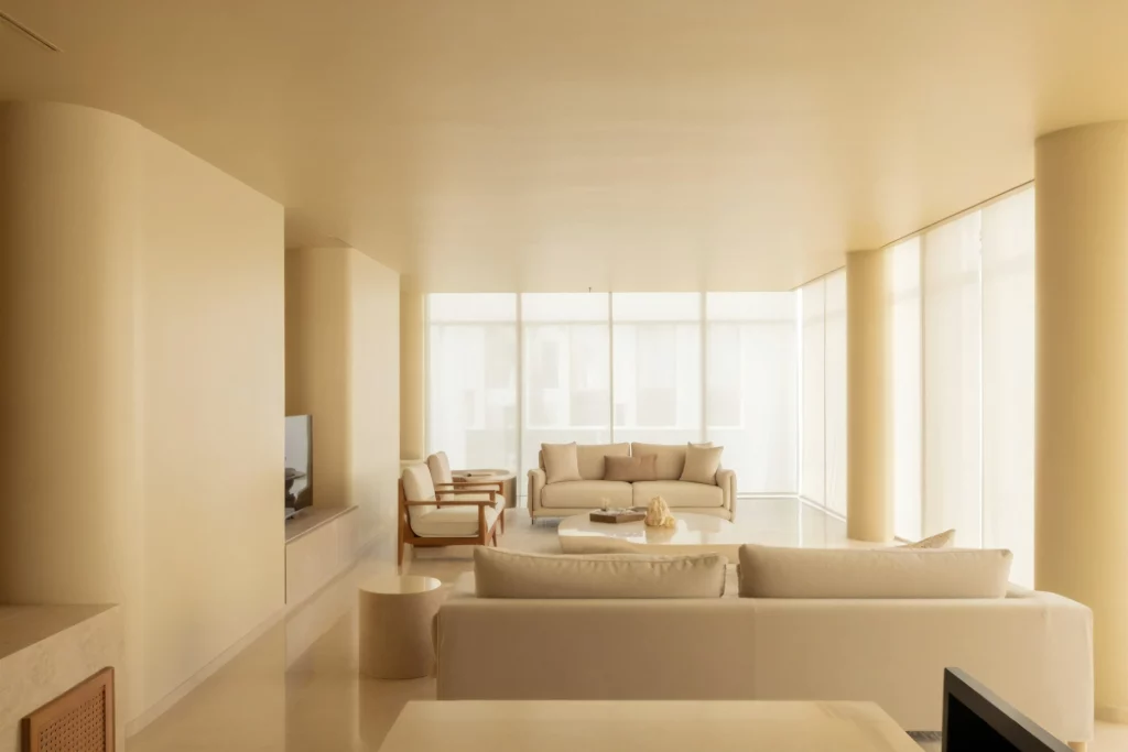 A minimalist living room with neutral tones, featuring clean lines, soft lighting, and modern furniture including a sofa, armchair, and round coffee table. Large windows allow natural light to fill the space.