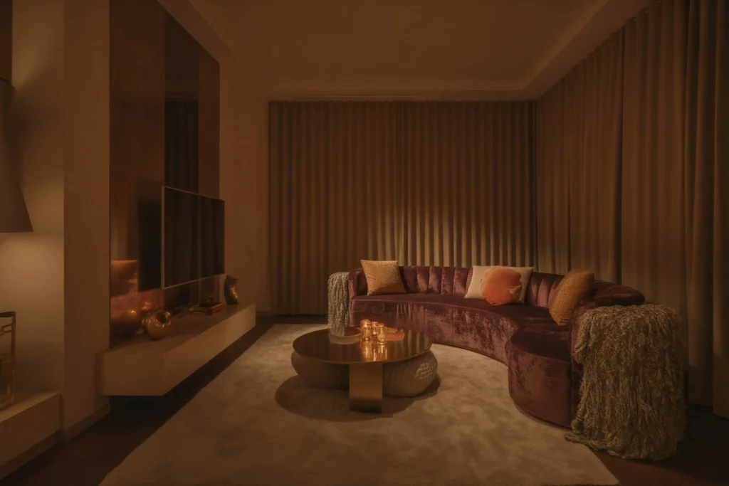 A cozy, dimly lit living room with a curved purple sofa adorned with colorful cushions. The space features a round coffee table with candles, warm lighting, and a minimalist design, creating an intimate and inviting atmosphere.