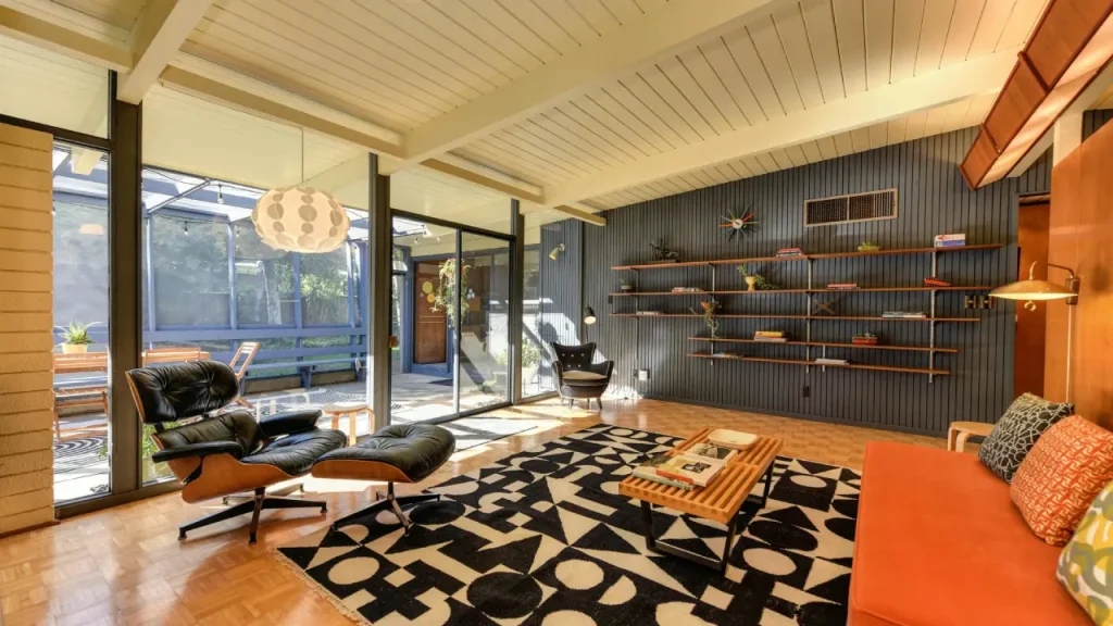 Mid-century modern living room with clean lines and functional furniture. The room features iconic Eames lounge chairs with sleek wooden frames, a low-profile orange sofa, and a geometric black and white rug. A wall of open shelving holds decorative items and plants, while large windows offer a view of an outdoor space. The room is flooded with natural light, highlighting the wooden parquet flooring and the retro pendant lamp hanging from the ceiling. The design combines timeless modern aesthetics with warm, earthy tones for a cozy yet stylish vibe.