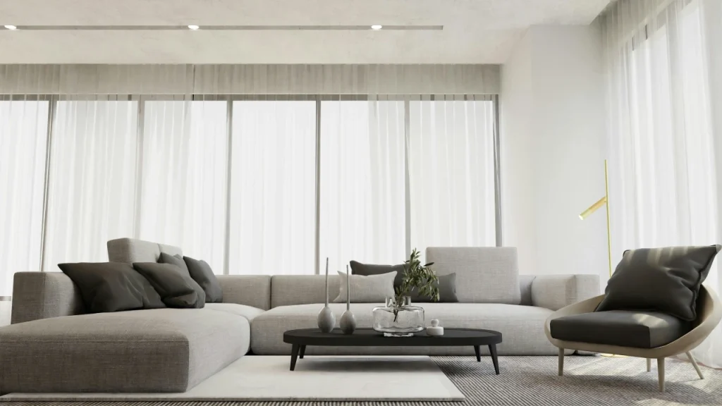 Modern minimalist living room featuring clean lines and a neutral color palette. The room is furnished with a spacious, light grey sectional sofa with dark grey pillows, a round black coffee table with a vase and candles, and a single light grey armchair. The space has large windows with sheer white curtains, allowing ample natural light. The flooring is a simple patterned rug, and the walls and ceiling have a smooth, neutral finish, creating a calm, uncluttered atmosphere. The design emphasizes simplicity, elegance, and functionality.